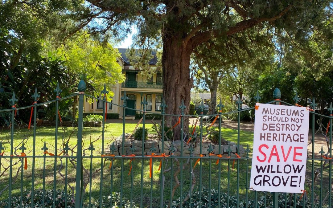 In the foreground, there is a fence with orange construction ties and a placard taped to it. The placard reads "Museums should not destroy heritage. Save Willow Grove". Behind the fence there is a large tree and behind it is the 1880s Italianate villa Willow Grove.