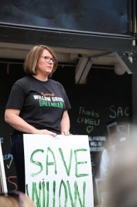 Councillor Donna Davis holds a placard. It reads "Save Willow Grove". Her hair is in a bob, she wears glasses and her shirt says "Save Willow Grove Greenban".